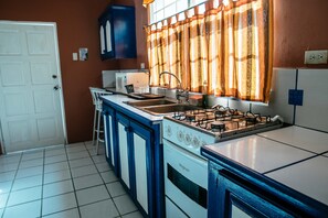 A kitchen with a white stove, blue cabinets, and a window with orange curtains. - Paradise Home Apartments (Canaan)