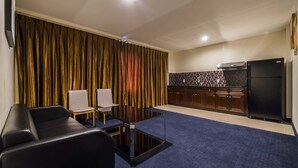 A modern living room with a black leather sofa, a glass coffee table, and two chairs. A kitchen with dark wooden cabinets and a black refrigerator is visible in the background.