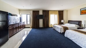 A hotel room with a large bed, a smaller bed, a desk with a chair, a flat-screen TV, a minibar, and a large window with curtains.