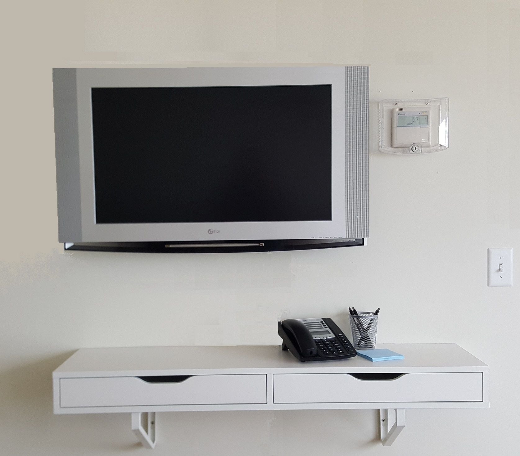 A flat-screen TV mounted on a wall above a white desk with a telephone and a pen holder.