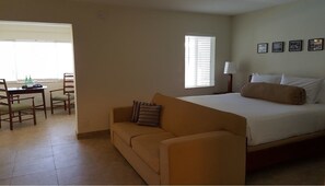 A hotel room with a bed, a sofa, a dining table with chairs, and a window with blinds. - Le Cabanon Motel (Pompano Beach)