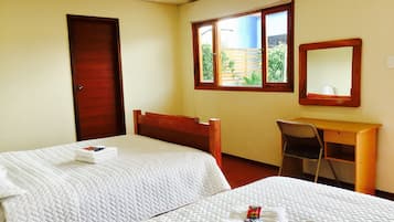 A hotel room with two beds, a desk, a chair, a window with a view of greenery, and a mirror on the wall.