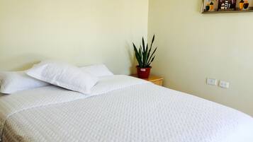 A neatly made bed with white bedding, a small potted plant on a bedside table, and a wall-mounted power outlet.
