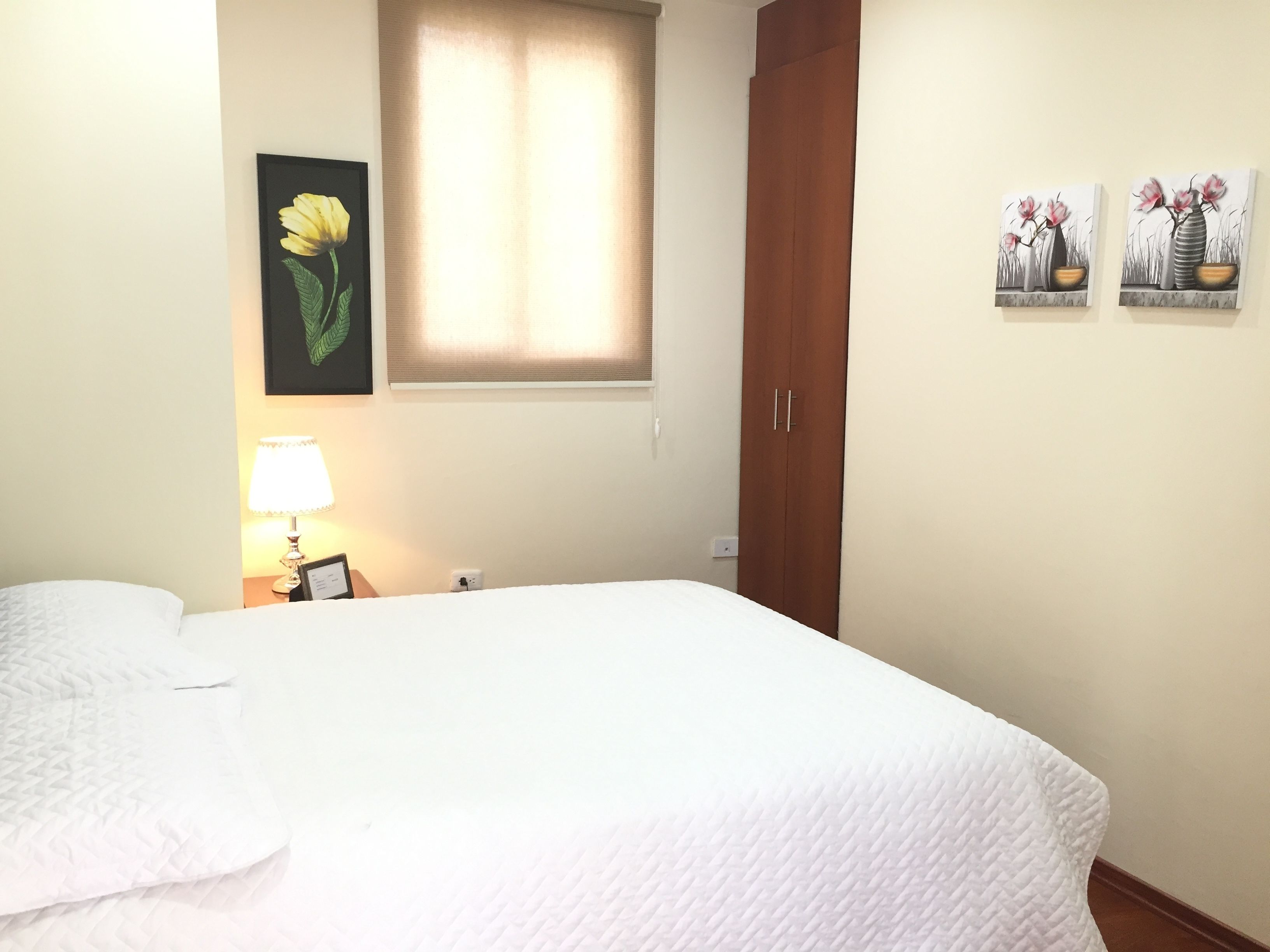 A neatly made bed in a room with a window, a lamp, and two wall-mounted paintings.