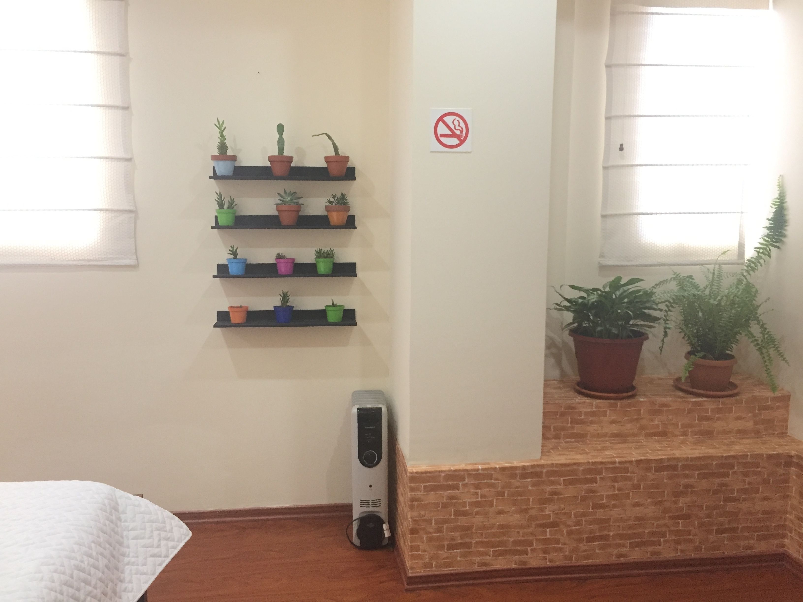 Ein Zimmer mit Ziegelwand, einem weißen Bett, einem an der Wand befestigten Regal mit Blumentöpfen, einem Rauchverbot-Schild und einem Fenster mit Jalousien.