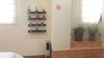 Ein Zimmer mit Ziegelwand, einem weißen Bett, einem an der Wand befestigten Regal mit Blumentöpfen, einem Rauchverbot-Schild und einem Fenster mit Jalousien.