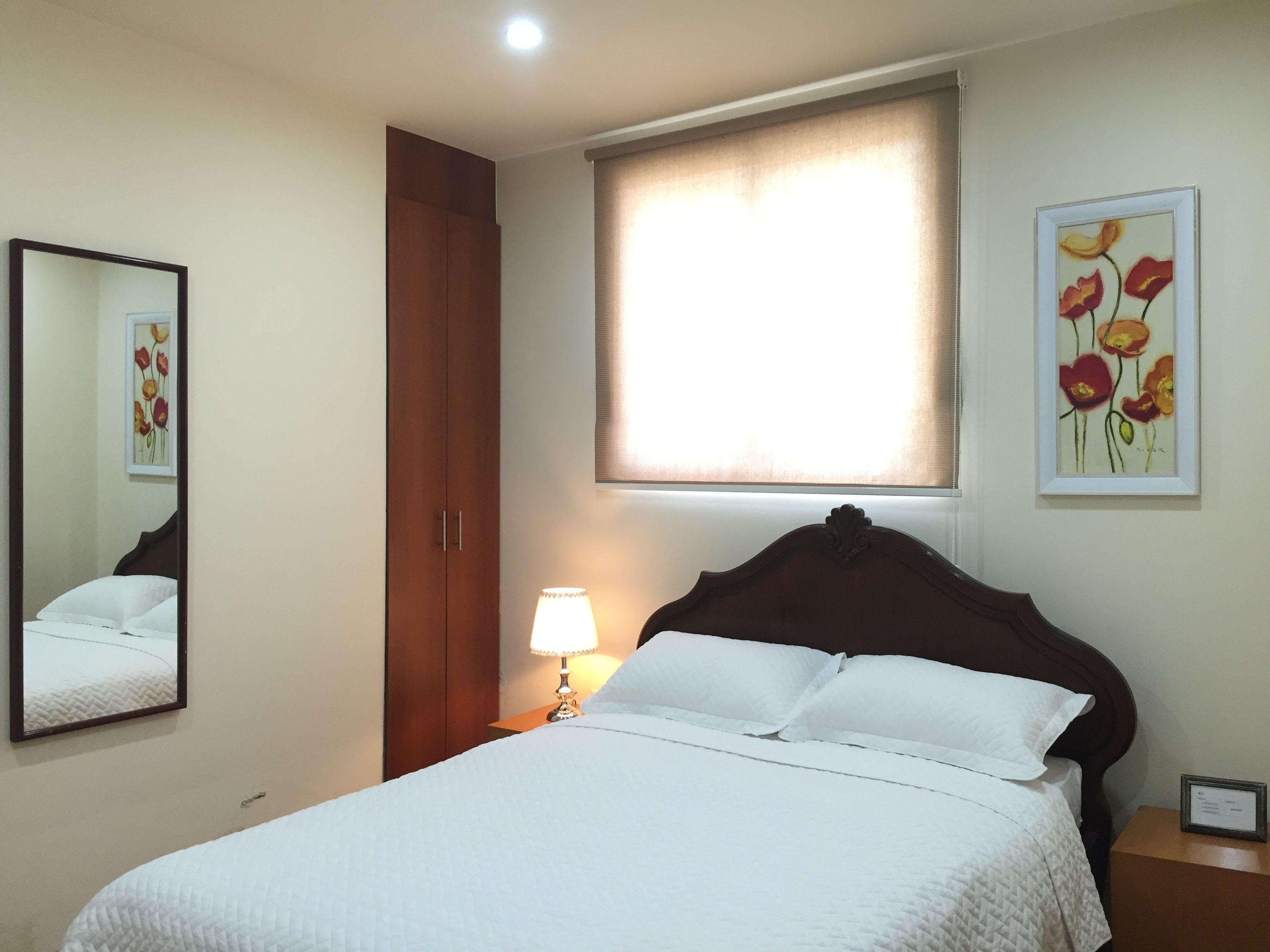 A hotel room with a bed, a nightstand with a lamp, a wardrobe, a window with blinds, and a framed floral artwork on the wall.