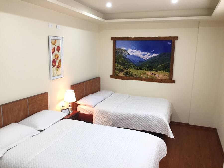 Habitación de hotel con dos camas, una mesita de noche con lámpara, un cuadro floral enmarcado y un cuadro de paisaje grande en la pared.