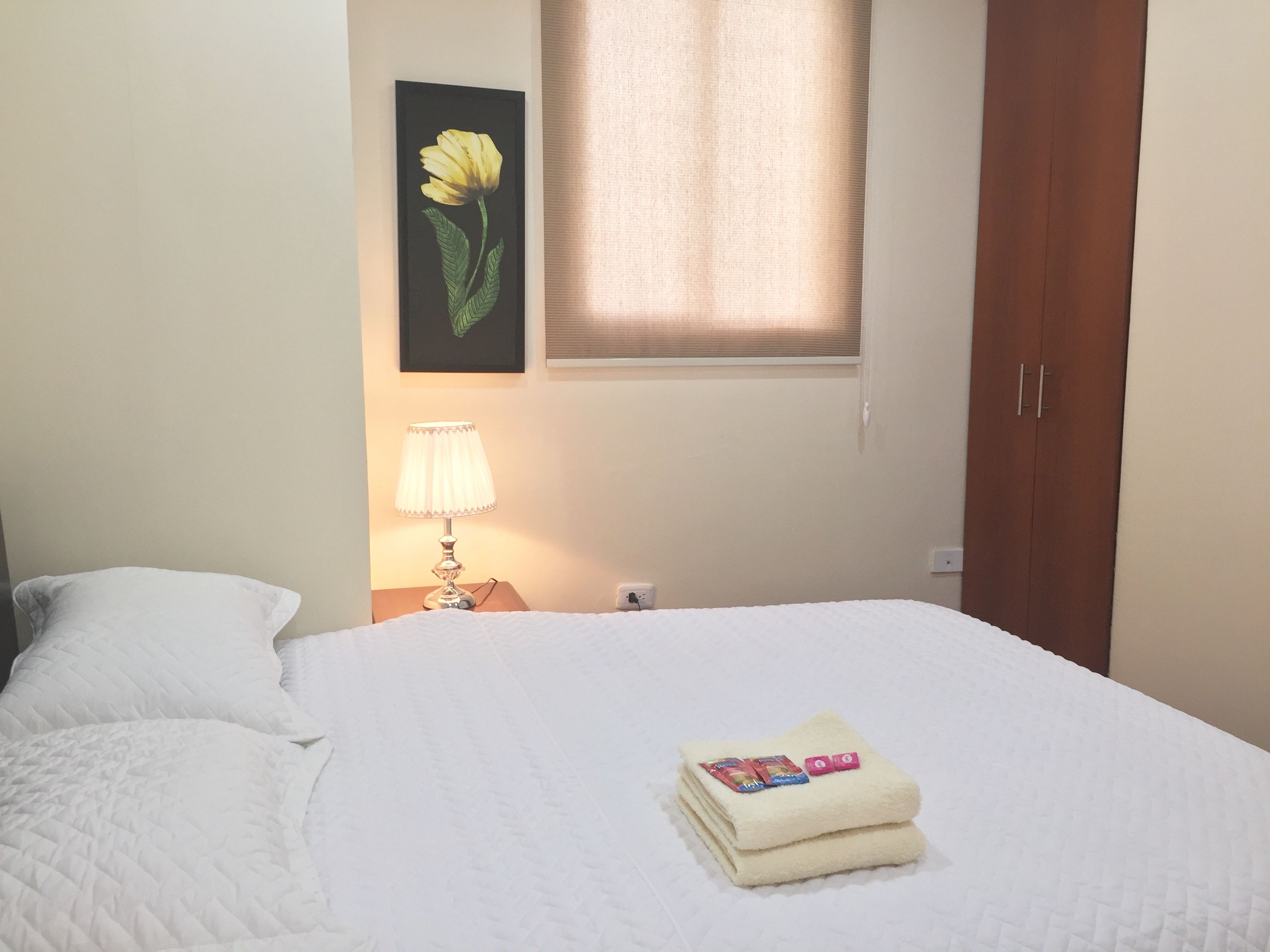 Una cama bien hecha con una toalla doblada y artículos de aseo, una lámpara de noche, un cuadro en la pared y una ventana con cortinas.