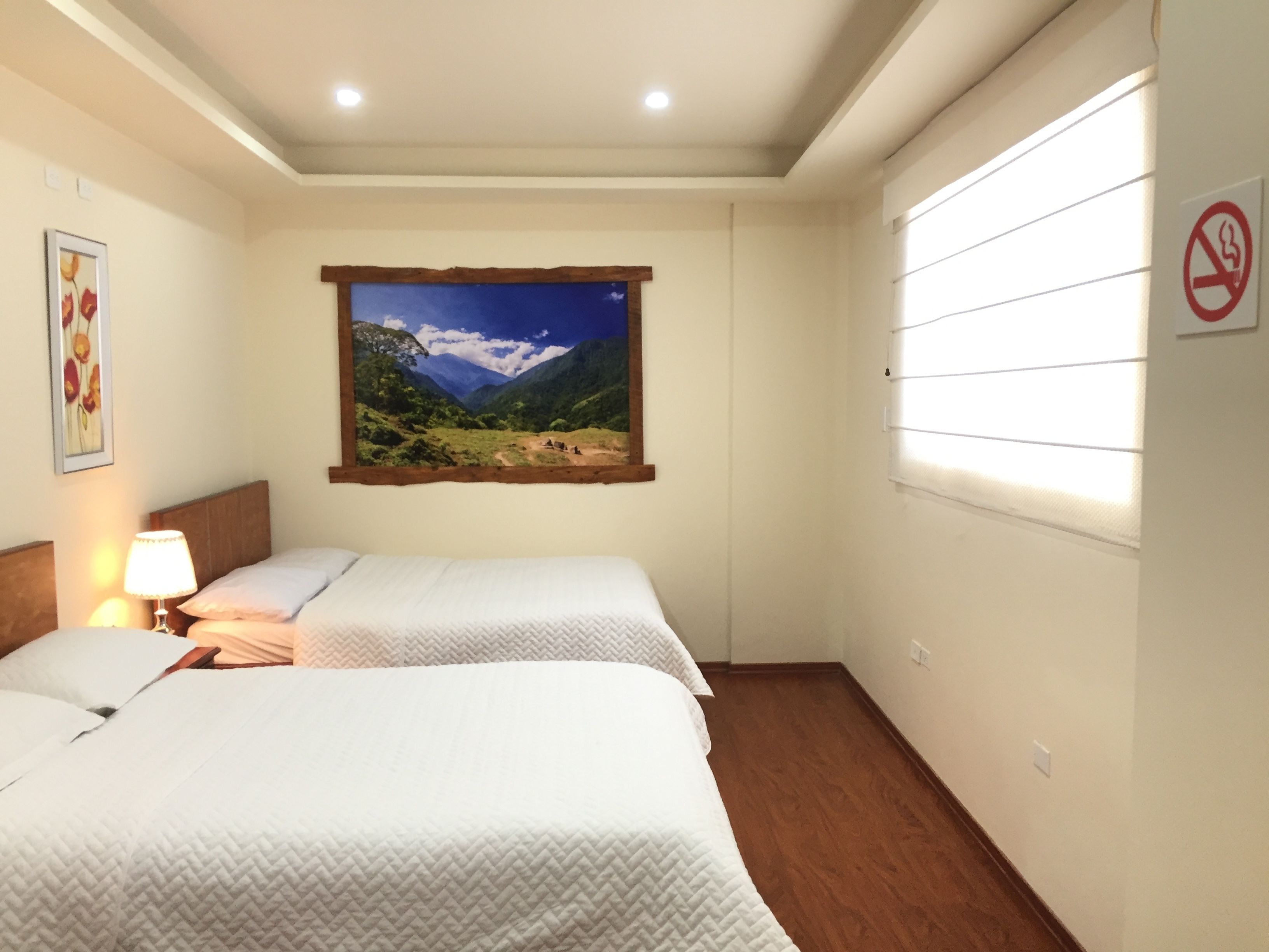 A hotel room with two beds, a painting of a mountainous landscape, a lamp, and a no-smoking sign.