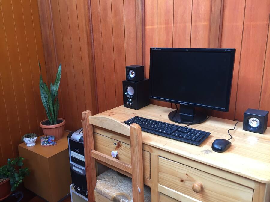 Un bureau en bois avec un ordinateur comprenant un écran, un clavier, une souris et des haut-parleurs. On y trouve également une chaise, une plante en pot et un petit meuble avec un distributeur d’eau.