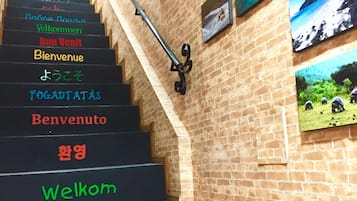 Escadaria com placas de boas-vindas em vários idiomas, corrimão de metal e parede de tijolos decorada com quadros.