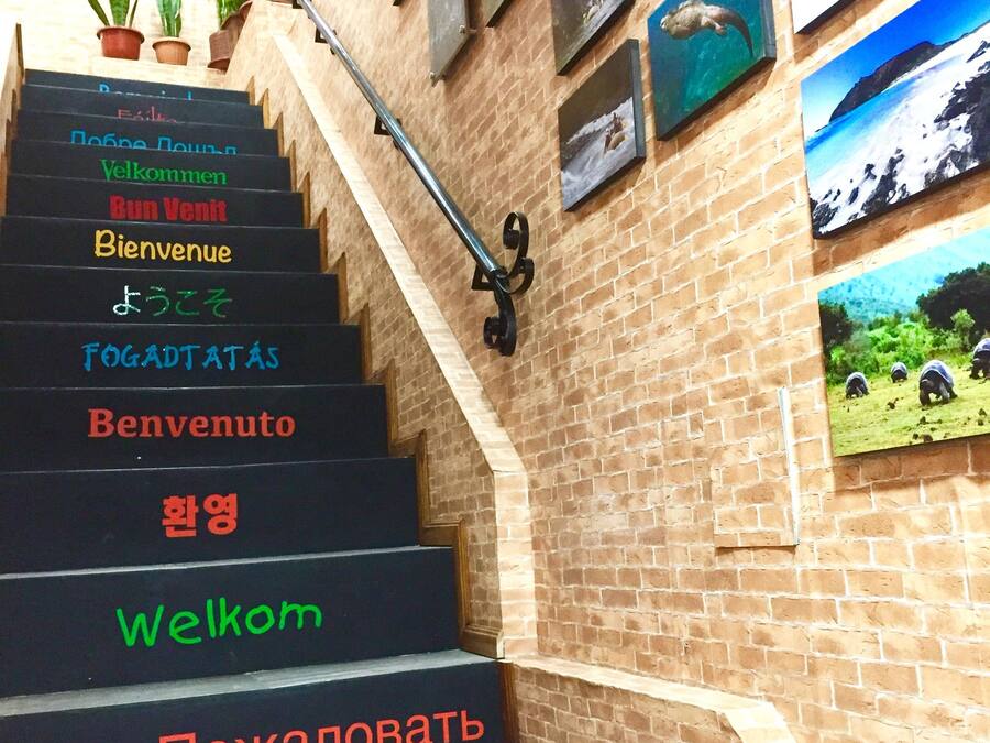 Escadaria com placas de boas-vindas em vários idiomas, corrimão de metal e parede de tijolos decorada com quadros.