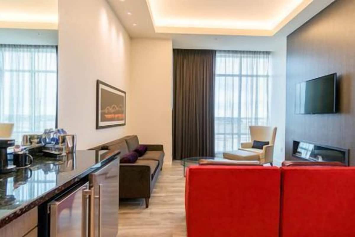 Una habitación de hotel moderna con comedor, sofá, un reposapiés rojo, televisor y ventanas amplias.