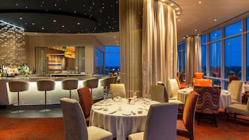 A modern dining area with round tables and bar seating, large windows, and a view of the cityscape.