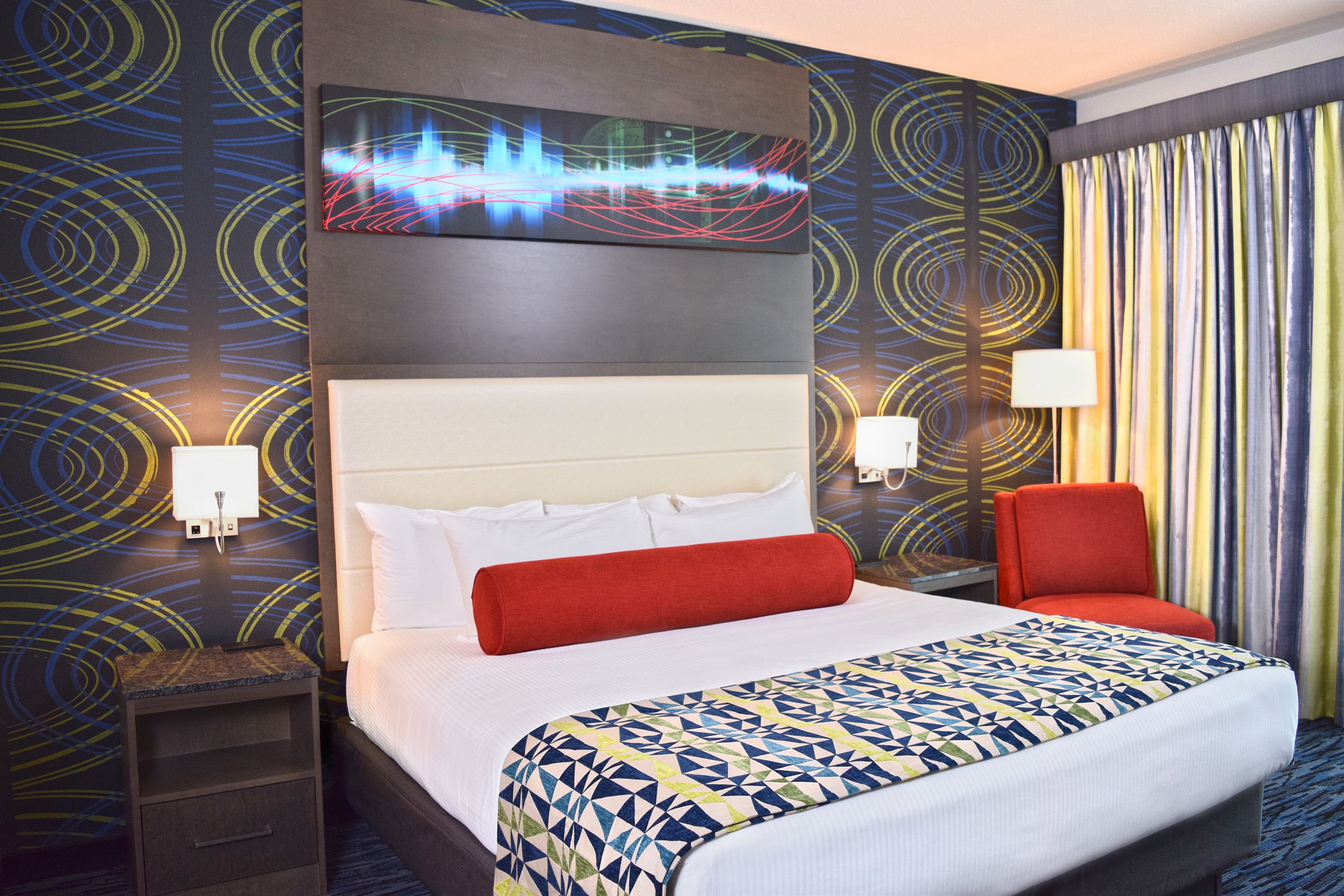 A hotel room with a large bed, a headboard, a nightstand, a red chair, and a wall with a colorful abstract design.