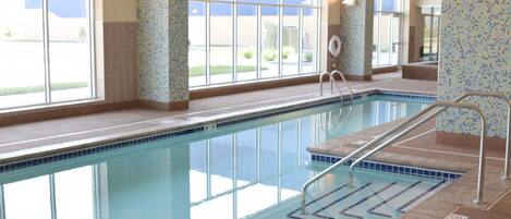 Una piscina interior con paredes de vidrio transparente, barandales de metal y suelo de baldosas.