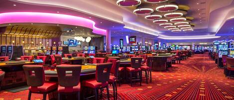 A casino with multiple gaming tables and chairs, a patterned carpet, and circular ceiling lights.