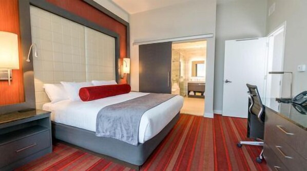 Habitación de hotel con cama, escritorio, silla y baño visible a través de una puerta abierta.
