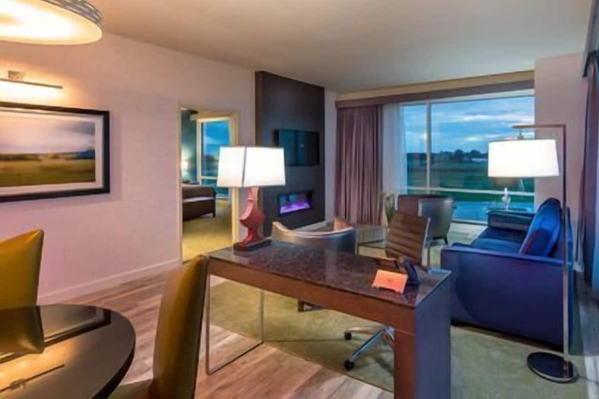 Habitación de hotel con comedor, sofá, una silla, lámpara y un gran ventanal con vistas.
