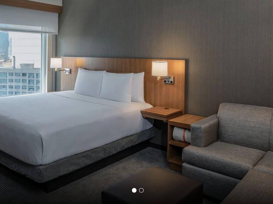A hotel room with a large bed, a grey sofa, a small table, and two wall-mounted lamps.