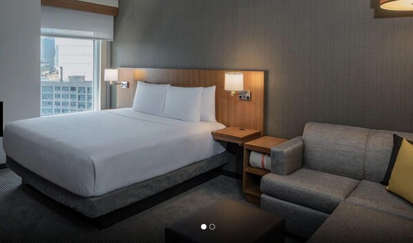 A hotel room with a large bed, a grey sofa, a small table, and two wall-mounted lamps.