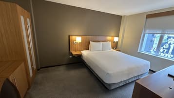 A hotel room with a large bed, two bedside tables with lamps, a wooden wardrobe, and a window with a view of the city.