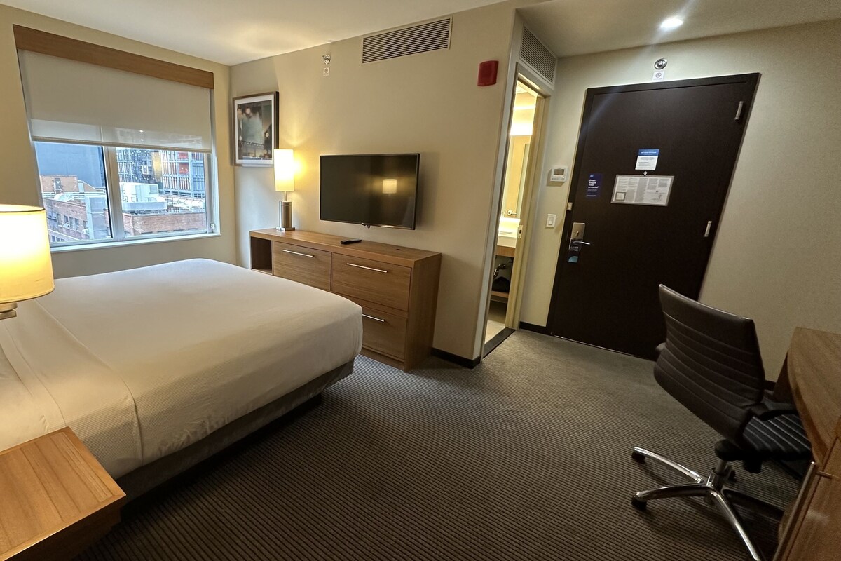 A hotel room with a bed, a TV, a desk, a chair, and a window with a city view.