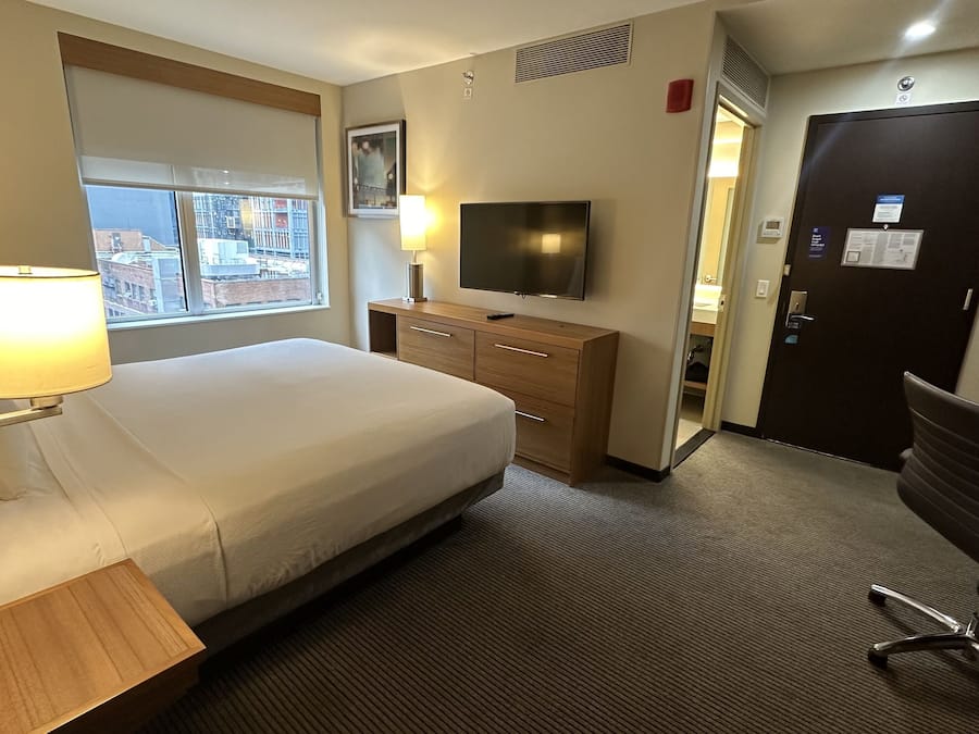 Habitación de hotel con cama, televisión, escritorio, silla y ventana con vista a la ciudad.