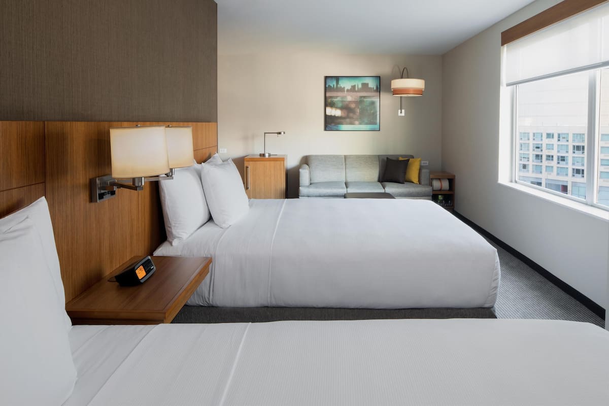Ein Hotelzimmer mit zwei Betten, einem Nachttisch mit Telefon, einer Lampe, einem Bild an der Wand und einem Fenster mit Blick auf die Stadt.