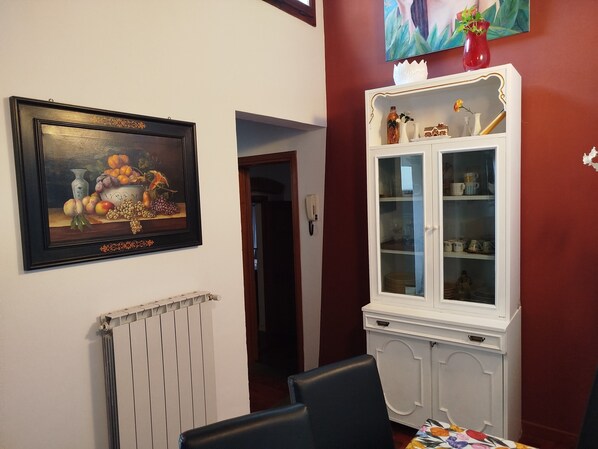 Uma área de refeições com uma mesa posta para dois, um armário branco com portas de vidro e uma pintura de natureza morta emoldurada na parede.