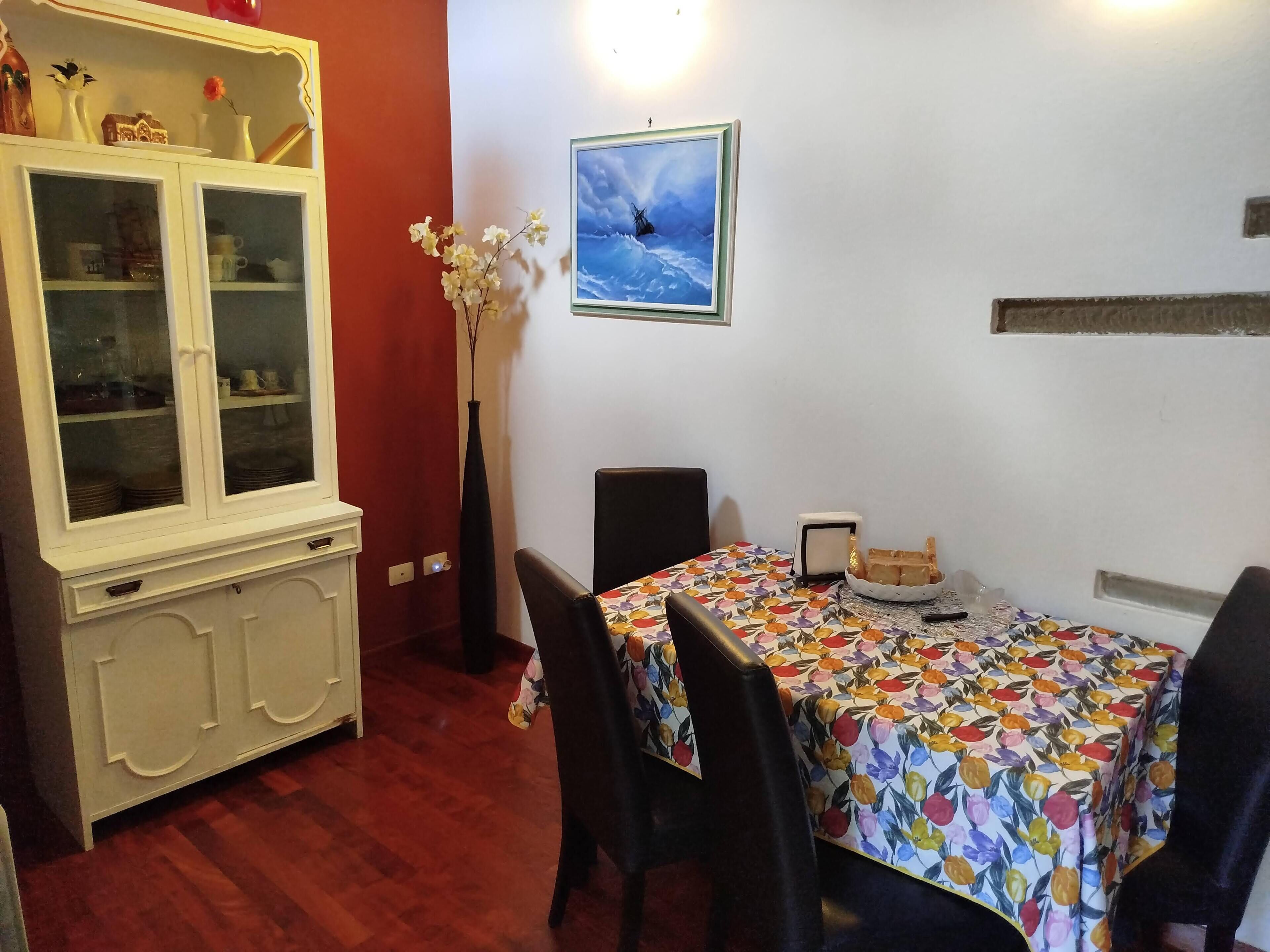 Un coin repas avec une table recouverte d’une nappe à motifs floraux, entourée de chaises. On aperçoit, à gauche, un meuble avec des portes en verre, et un vase rempli de fleurs est posé sur le sol, à côté de la table.