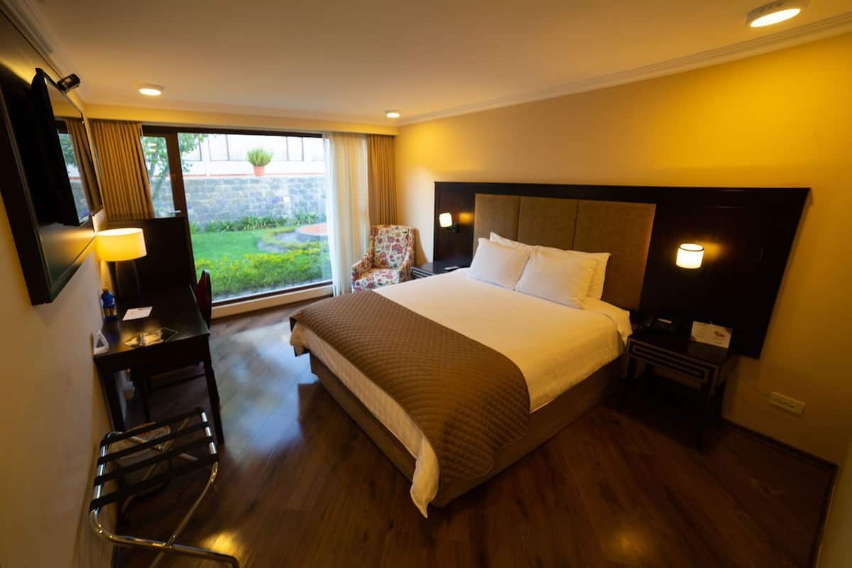A hotel room with a large bed, a desk with a lamp, a chair, and a view of a garden through a window.