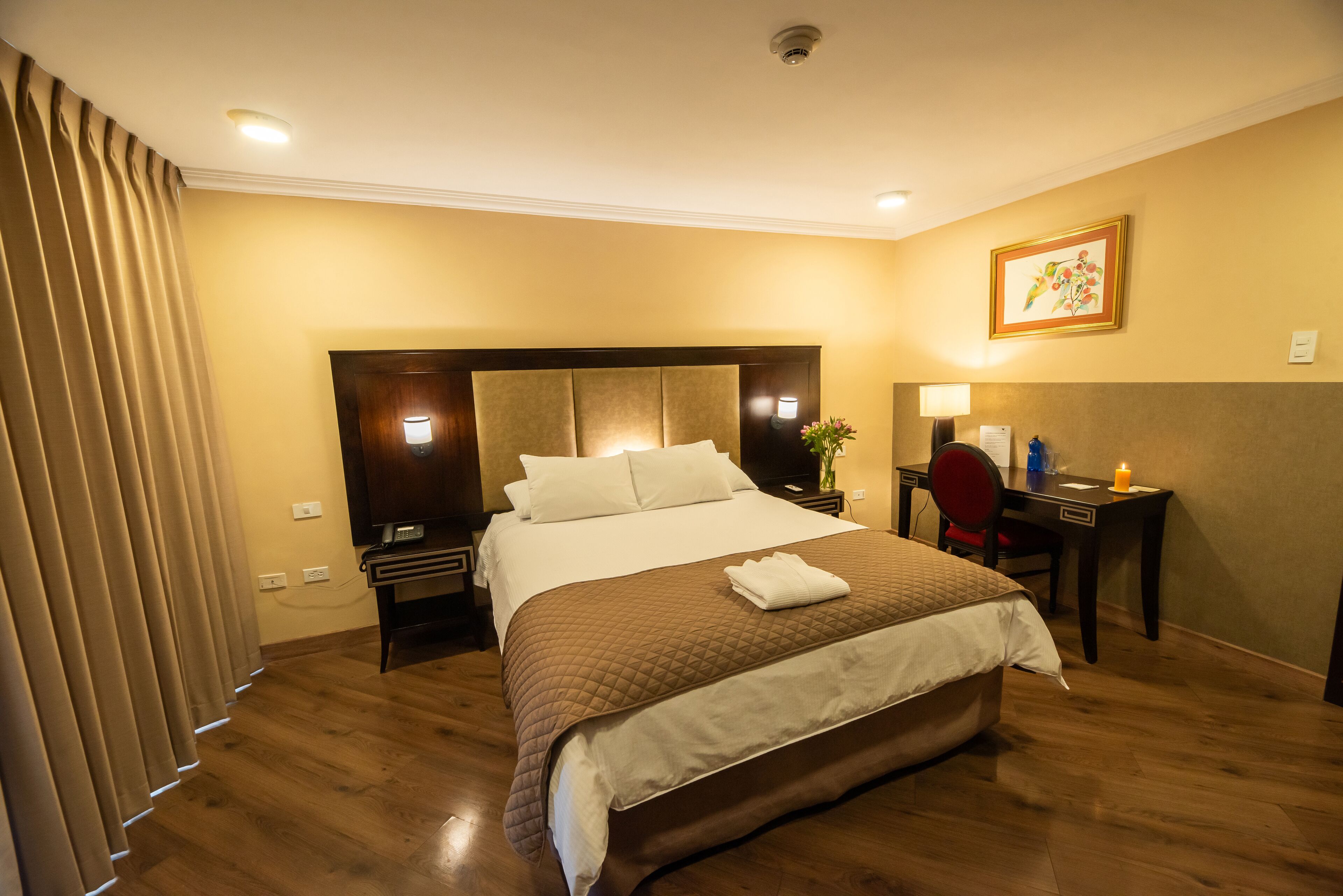 Une chambre d’hôtel avec un grand lit, un bureau et une chaise. On y trouve une table de chevet avec une lampe et un vase de fleurs. La chambre est dotée d’un parquet et de murs de couleur beige.