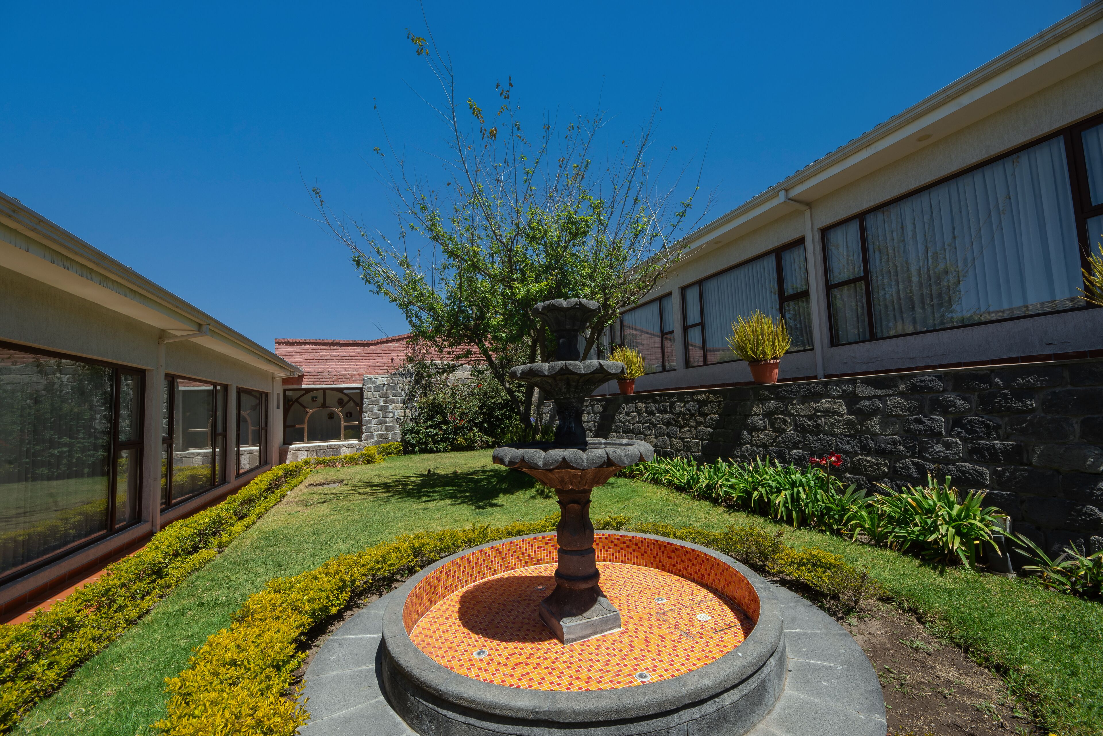 Un patio con una fuente de piedra, un césped bien cuidado y un edificio con grandes ventanales y un muro de piedra.