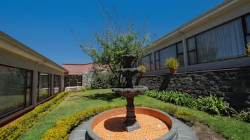 Un patio con una fuente de piedra, un césped bien cuidado y un edificio con grandes ventanales y un muro de piedra.