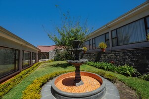 Un patio con una fuente de piedra, un césped bien cuidado y un edificio con grandes ventanales y un muro de piedra.