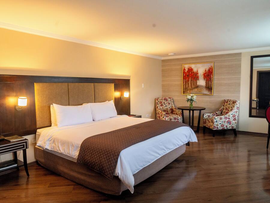 A hotel room with a large bed, bedside tables, a desk, a chair, and a small table with a vase.