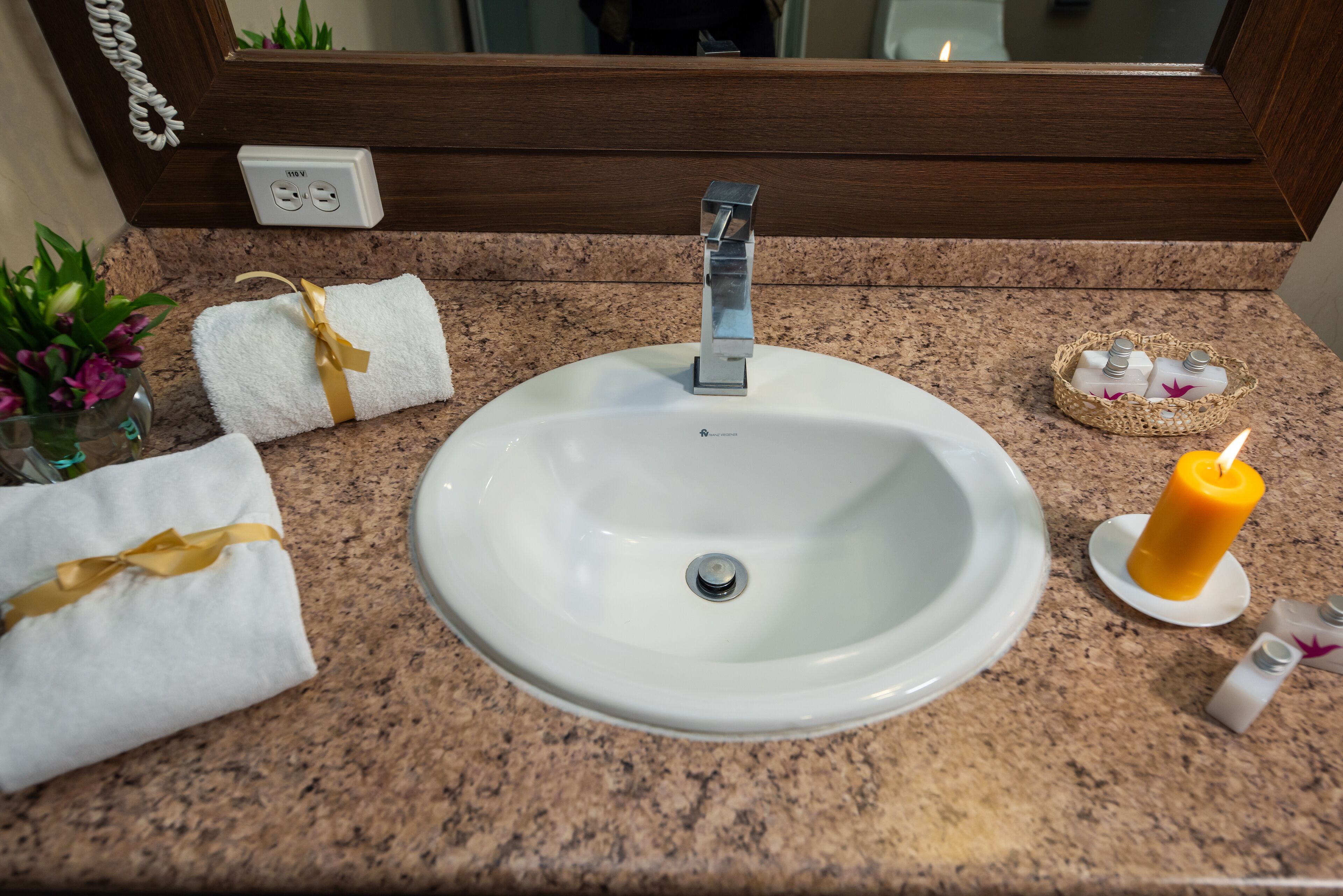 A bathroom sink with a mirror above it, a towel with a yellow ribbon, a candle, and toiletries on the countertop.