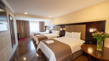 Habitación de hotel con tres camas, piso de madera, un espejo grande, mesita de noche con lámpara y un jarrón con flores.