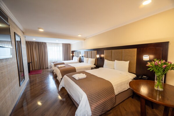 Habitación de hotel con tres camas, piso de madera, un espejo grande, mesita de noche con lámpara y un jarrón con flores.