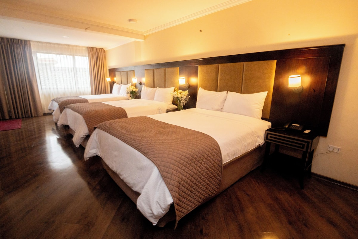 A hotel room with three beds, wooden flooring, and a nightstand with a lamp.