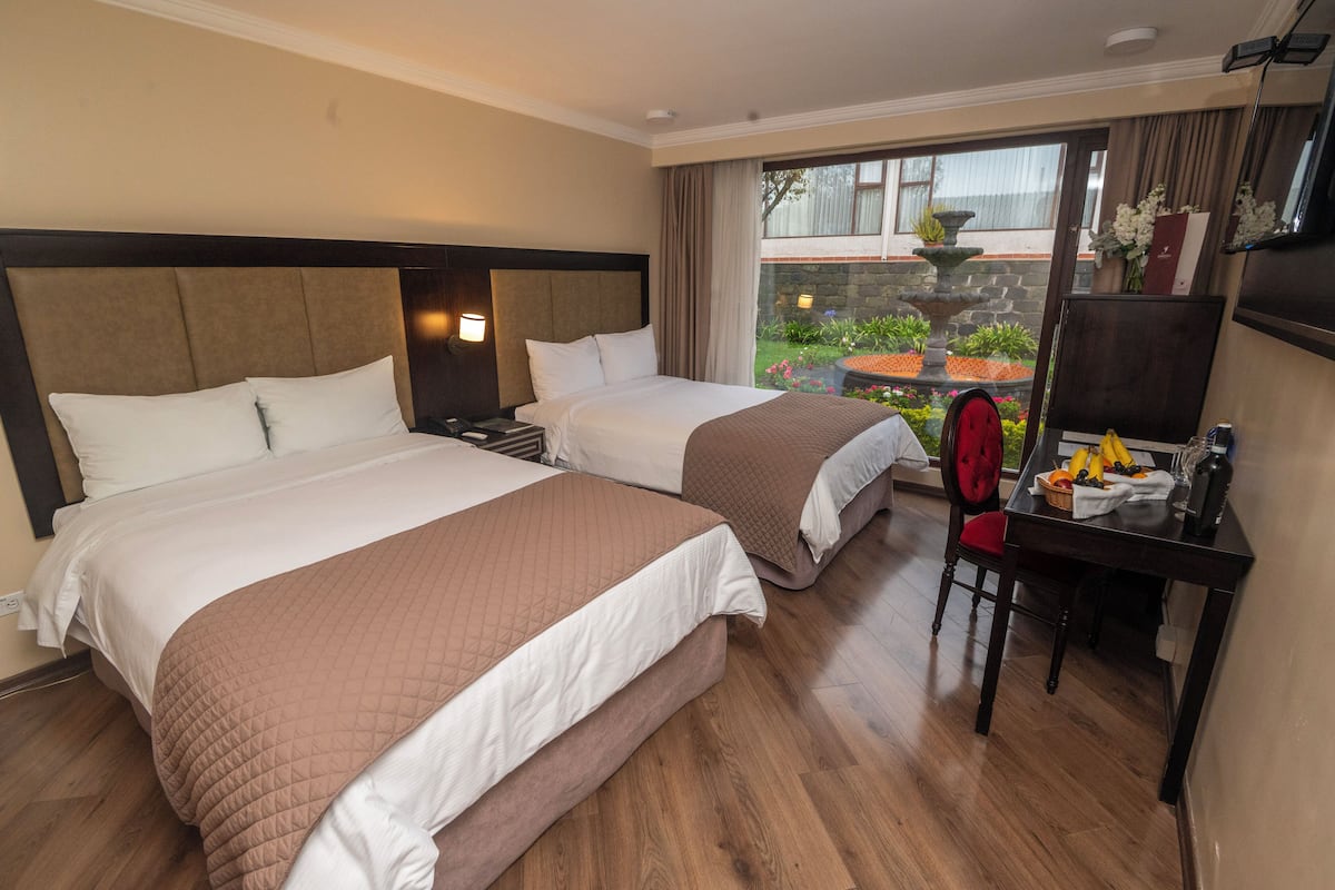 A hotel room with two beds, a desk with a chair, and a view of a garden through a glass door.