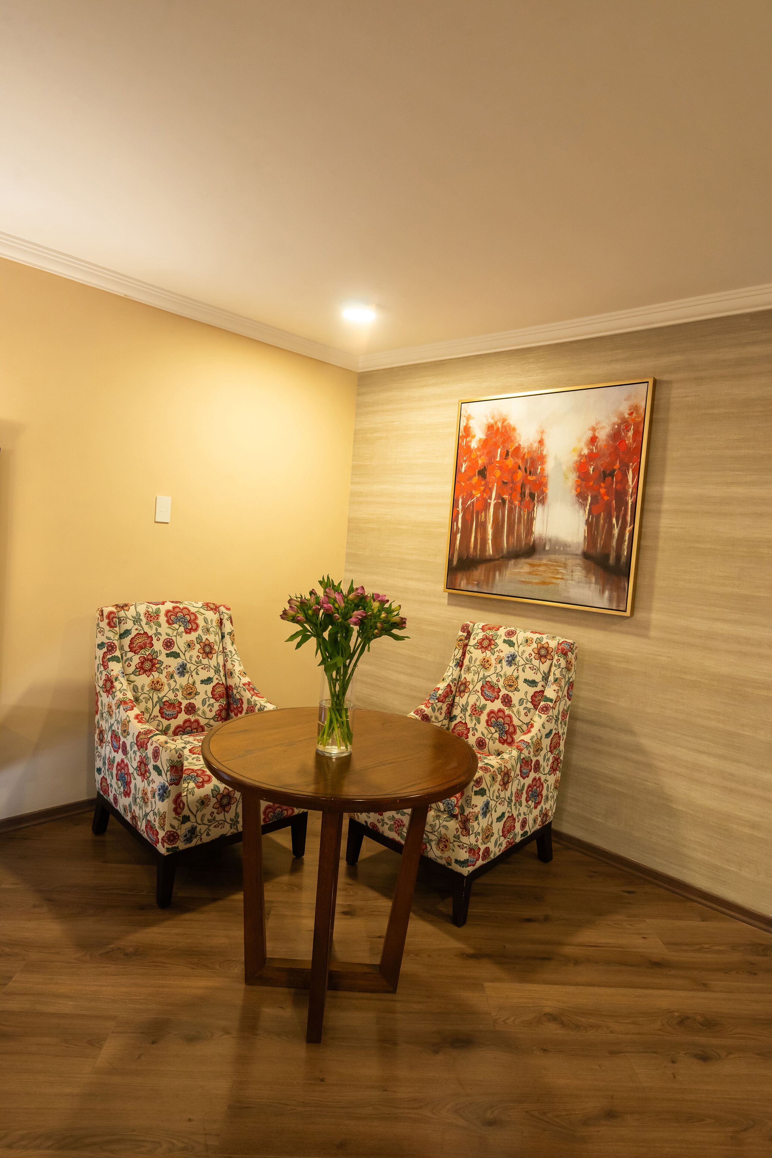 Una habitación con dos sillas de estampado floral, una mesa de madera y un cuadro de árboles otoñales en la pared.