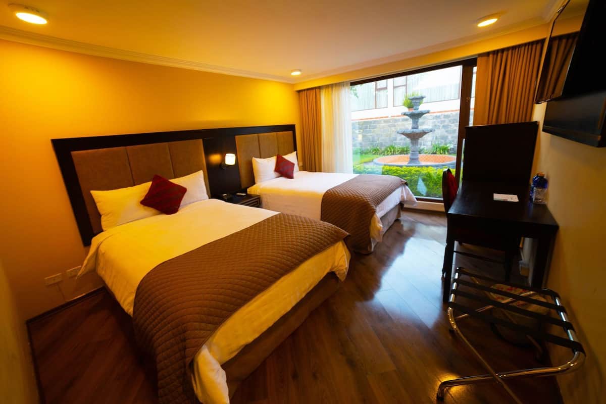 A hotel room with two beds, a TV, a desk, and a view of a garden.