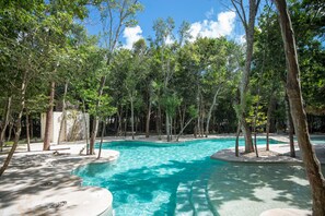 Une piscine aux eaux cristallines, entourée d’arbres, avec un mur de pierre et une zone sablonneuse à proximité.