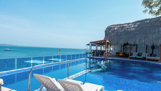 A clear blue pool with white lounge chairs, a thatched-roof structure, and a view of the sea. - Hotel Isla Real (Cartagena)