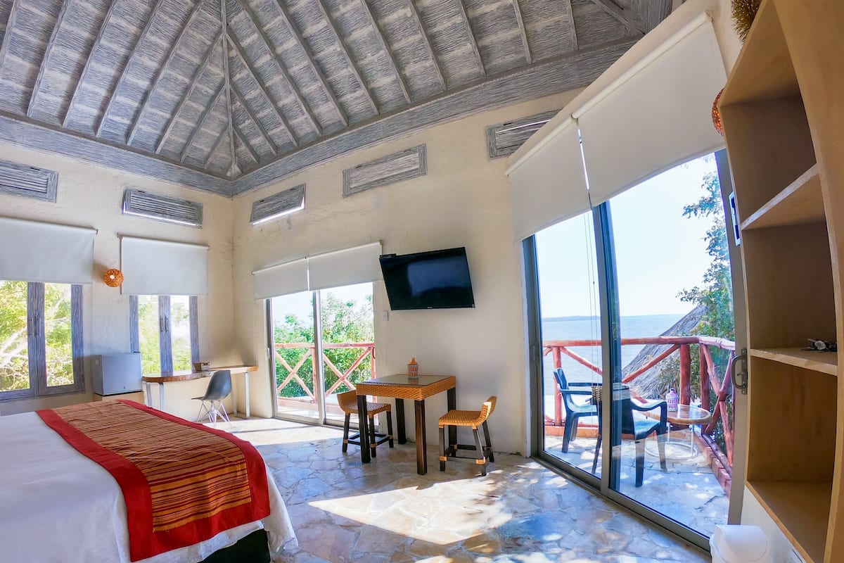 Una habitación con cama, televisión, mesa con sillas, balcón con barandilla y vista al mar.