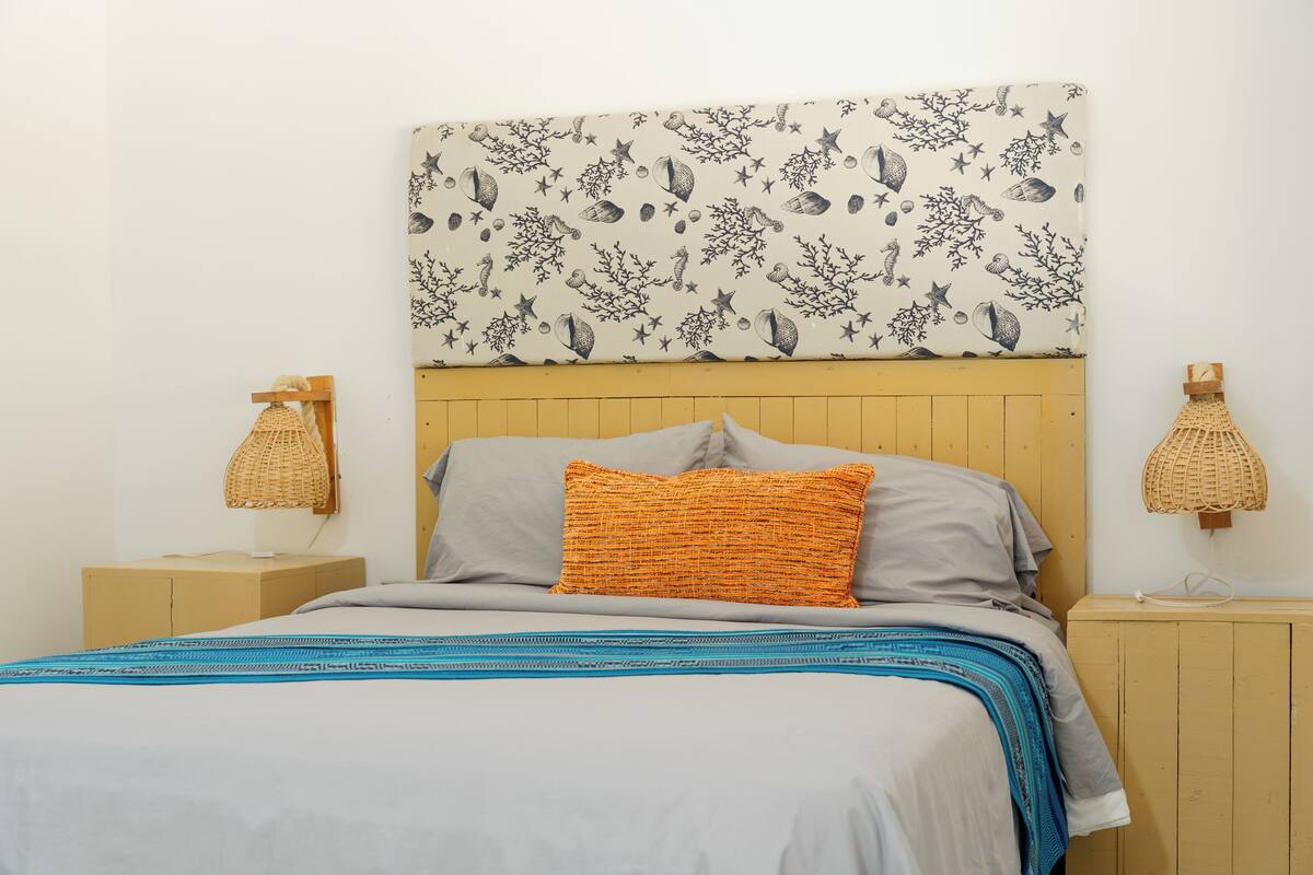 Una cama con un cabecero estampado, un almohadón gris, un almohadón naranja y una colcha azul y blanca con motivos.