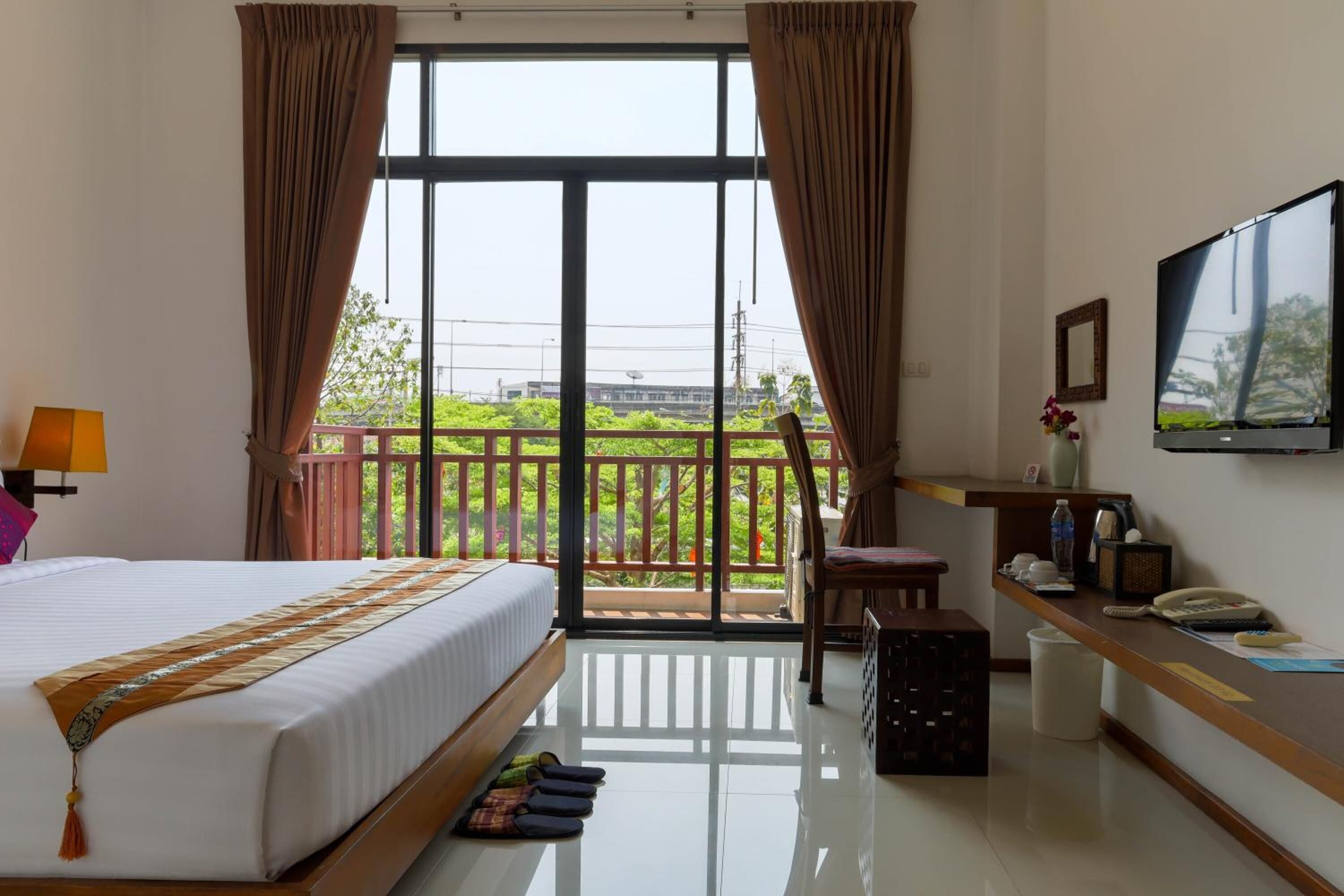 A hotel room with a bed, a desk, a chair, a TV, and a balcony with a view of greenery.